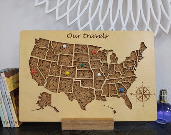 May include: A wooden map of the United States with a cork surface. The map has a compass in the bottom right corner and the words "Our travels" at the top. There are colorful push pins marking different locations on the map.