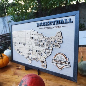 Può includere: Una mappa in legno degli Stati Uniti a tema basket. La mappa è incorniciata in nero e ha un supporto in legno marrone. La mappa ha dei fori per le puntine e il testo "Basketball Stadium Map" è in alto. La parte inferiore della mappa ha un logo in legno marrone con il testo "Miller Family Stadium Map" e una grafica di basket.