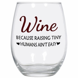 May include: A clear glass wine glass with the text "Wine because raising tiny humans ain't easy" printed in maroon on the front.