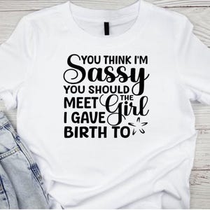 以下が含まれることがあります： 「YOU THINK I'M SASSY YOU SHOULD MEET THE GIRL I GAVE BIRTH TO」という黒い文字がプリントされた白いTシャツ。青いデニムのジーンズと組み合わせています。
