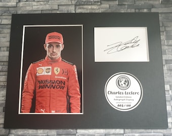 May include: A black framed photo of Charles Leclerc, a Formula 1 driver, wearing a red racing suit with the Ferrari logo and the words "Mission Winnow" on it. The photo is signed by Leclerc and includes a limited edition autograph display with the number 002/100.
