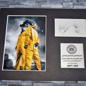 May include: Framed print with two figures in yellow hazmat suits, set against a smoky backdrop. Includes a signed card and a limited edition certificate. The certificate reads "Limited Edition Autograph Display" and the edition number is 007/100.