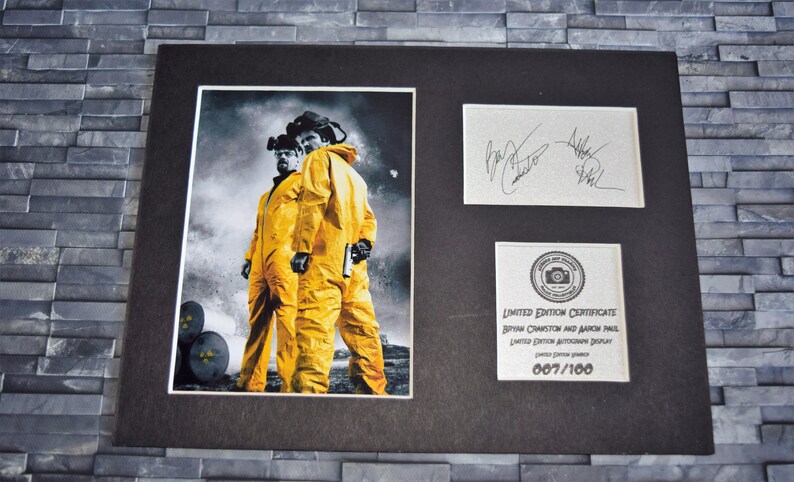 May include: Framed print with two figures in yellow hazmat suits, set against a smoky backdrop. Includes a signed card and a limited edition certificate. The certificate reads "Limited Edition Autograph Display" and the edition number is 007/100.