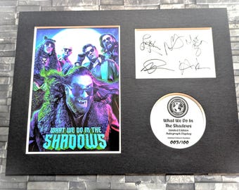 May include: A framed, signed poster of the cast of the TV show "What We Do in the Shadows". The poster features a colourful image of the cast members, with the show's title in a stylised font. The poster is numbered 003/100, indicating it is part of a limited edition.