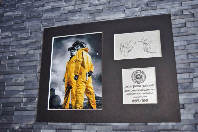 May include: Framed print depicting two figures in yellow hazmat suits, set against a grayscale backdrop with a nuclear symbol. Includes signed cards and a limited edition certificate numbered 007/100.