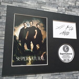 May include: A framed photograph of the three main actors from the TV show Supernatural, with their autographs. The photo is black and white and shows the actors standing in a field. The frame is black and has a white mat. The photo is signed by Jared Padalecki, Jensen Ackles, and Misha Collins. The frame also includes a round, white sticker with the text "Supernatural Limited Edition Autograph Display 003/100".