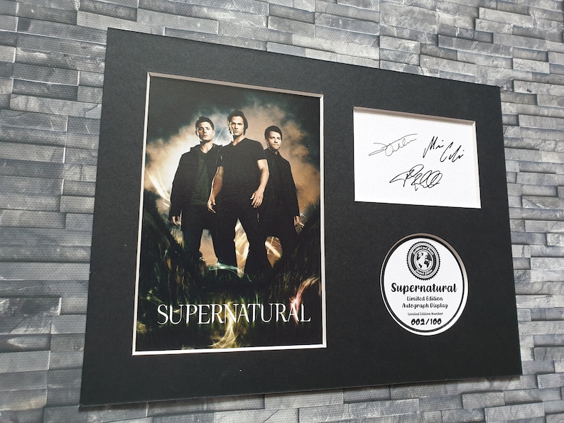 May include: A framed photograph of the three main actors from the TV show Supernatural, with their autographs. The photo is black and white and shows the actors standing in a field. The frame is black and has a white mat. The photo is signed by Jared Padalecki, Jensen Ackles, and Misha Collins. The frame also includes a round, white sticker with the text "Supernatural Limited Edition Autograph Display 003/100".