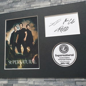 May include: A black framed print of a photo of three men from the TV show Supernatural. The print has a black and white signature, a Supernatural logo, and the text "Limited Edition Autograph Display 002/100".