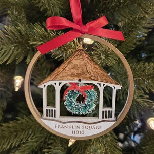 May include: A gold-colored round ornament with a white gazebo and a green wreath inside. The ornament has a red ribbon and the text "Franklin Square 11010" on the bottom.