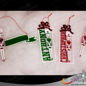 Puede incluir: Tres adornos navideños personalizados con un diseño de soldado de juguete. Los adornos son blancos con detalles rojos y verdes. Los nombres "Anthony", "Christopher" y una pancarta en blanco están impresos en los adornos.