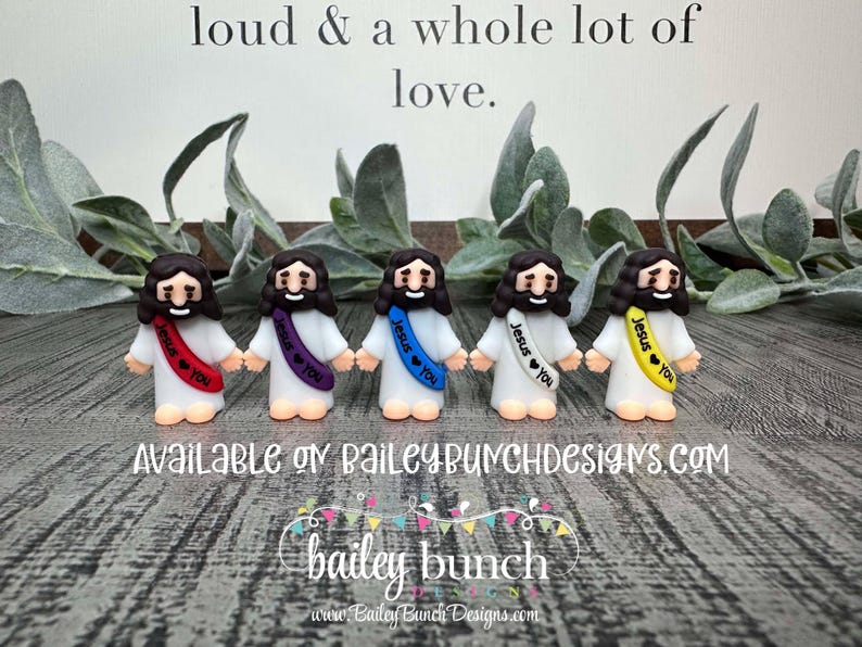 May include: Five small figurines of Jesus with different colored sashes that say "Jesus loves you" in white lettering. The figurines are standing on a wooden surface.
