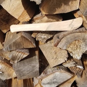 May include: A close-up of a wooden handle for a tool, possibly a hammer or axe. The handle is light brown and has a smooth surface. It is lying on top of a pile of stacked firewood.