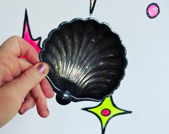Puede incluir: Un plato de plata con forma de concha marina con una superficie texturizada. El plato está siendo sostenido por una mano.
