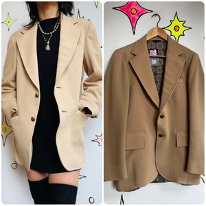 May include: A tan blazer with a two-button closure, notched lapels, and two front pockets. The blazer is shown on a hanger and worn by a person. The blazer has a patterned lining and is made of a textured fabric. The blazer is a neutral color.
