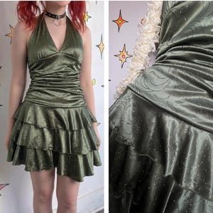 Puede incluir: Vestido halter verde oliva con falda escalonada. El vestido tiene cuello en V y corpiño entallado. La tela tiene un patrón sutil y un ligero brillo. El vestido es de largo mini.