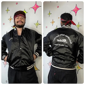 May include: Black satin bomber jacket with white and black striped trim. The front of the jacket has a cursive signature, and the back features the text "Pasadena Auto Auction" and a graphic of vehicles. The jacket has a button-up front and pockets.