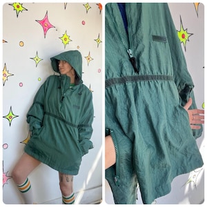 May include: A dark green anorak-style jacket with a hood, a half-zip closure, and a drawstring waist. The jacket has a small logo patch on the chest and two front pockets. The sleeves are long, and the jacket is made of a lightweight, crinkled fabric.
