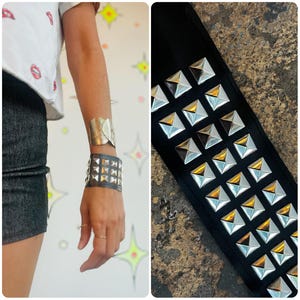 Puede incluir: Una pulsera de brazalete plateada y una pulsera negra con tachuelas se usan en la muñeca. La pulsera con tachuelas tiene tachuelas piramidales plateadas. También se muestra un cinturón negro con tachuelas con tachuelas piramidales plateadas y doradas.
