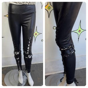 May include: Black faux leather leggings with lace-up detailing on the lower legs. The leggings have a high waist and are displayed on a mannequin. The design features cut-out sections with criss-cross lacing.
