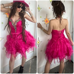 May include: A vibrant fuchsia-colored party dress with a layered, ruffled skirt. The bodice is embellished with sparkling gemstones. The dress features a halter neckline and a lace-up back. The dress is a fun and eye-catching choice for special occasions.
