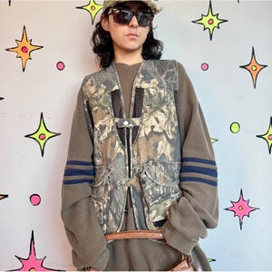 Puede incluir: Una sudadera marrón de manga larga con dos rayas azul oscuro en cada manga, superpuesta con un chaleco utilitario de estampado de camuflaje. El chaleco cuenta con paneles de malla negra, cierre de cremallera frontal y varios bolsillos con botones.