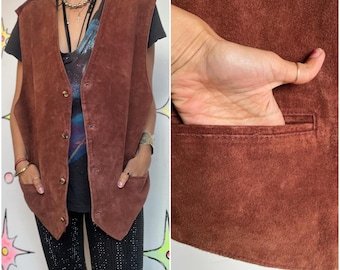 Puede incluir: Un chaleco de ante marrón con cuello en V, cierre de botones y dos bolsillos delanteros. El chaleco tiene un acento burdeos en los hombros. La imagen también muestra un primer plano del bolsillo del chaleco.