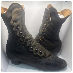 Puede incluir: Un par de botas negras antiguas con cierres de botones y bordes festoneados. Las botas tienen un tacón bajo y una estética vintage, mostrando un diseño clásico de una época pasada.