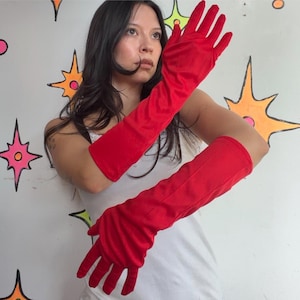 May include: A pair of long, solid red opera-length gloves made of a smooth, matte fabric. The gloves extend past the elbow, covering the forearms and hands completely with a fitted silhouette. The material appears opaque and uniform in color.
