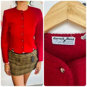 Puede incluir: Un cárdigan rojo brillante, de manga larga, con cuello redondo y botones decorativos. El dobladillo es festoneado. La etiqueta dice "Carroll Reed Ski Shops New Hampshire".