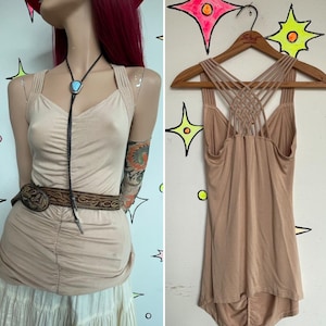 May include: A beige sleeveless tank top featuring a ruched front and a complex lattice of thin straps across the back. The top is shown on a mannequin with a bolo tie and a brown tooled leather belt, and also displayed on a wooden hanger.