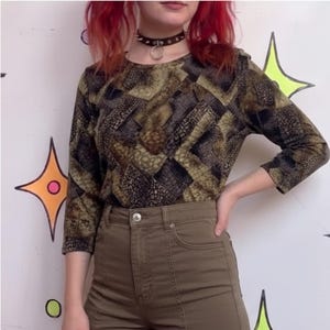 May include: A long-sleeve shirt featuring a dark brown, black, and olive green geometric snakeskin-like pattern. The shirt has a round neckline and is paired with high-waisted olive green pants that feature a central vertical seam and a silver button.