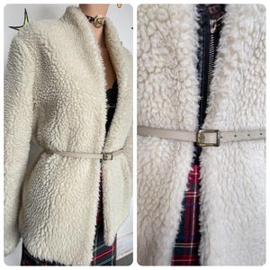 May include: Cream-colored, textured fleece jacket with a shawl collar and a matching belt. The jacket has a zip closure and a light-colored belt with a square buckle. The jacket is worn over a black top and a plaid skirt.