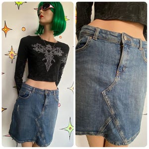 May include: A denim skirt with a unique angled design. The skirt is a medium wash blue with a distressed detail on the front. The skirt is paired with a black long-sleeve crop top with a silver design.