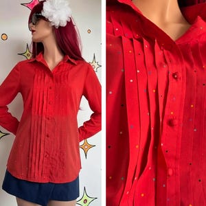 May include: A long-sleeved red button-down shirt featuring vertical pleats along the front placket and a pattern of small, multicolored polka dots. The shirt has a classic collar and matching red fabric-covered buttons down the center front.