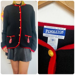 May include: Black Pendleton wool cardigan with red trim and gold buttons. The jacket features red pocket accents with red and white tassels. The label reads "Pendleton 100% Virgin Wool Portland Oregon".