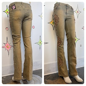 May include: Tan-colored denim jeans with decorative brown leather patches and stitching along the sides. The jeans have a flared leg and a faded appearance. A small tag is visible on the back pocket.