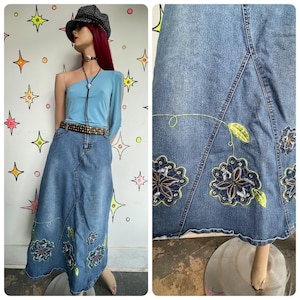 May include: A denim maxi skirt with floral embroidery in shades of blue, green, and yellow. The skirt is paired with a light blue one-shoulder top, a studded belt, and a patterned hat. The skirt features a flared design.
