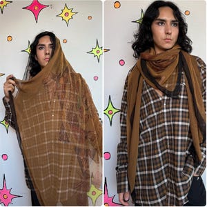 May include: A sheer brown scarf with a white grid pattern and a dark brown border, worn draped over the head and shoulders, paired with a brown, black, and white plaid long-sleeve button-down shirt featuring a collar and buttoned cuffs.