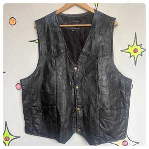 May include: Black leather vest with a patchwork design. The vest has a V-neck, four gold-colored buttons, and two front pockets. It is hanging on a wooden hanger against a wall with colorful star and circle designs.
