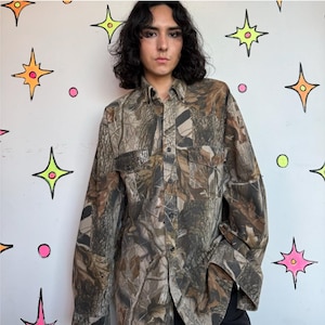 Puede incluir: Una camisa de manga larga con botones y un patrón de camuflaje forestal realista en tonos marrón, beige y verde oscuro. La camisa incluye un cuello estándar, una tapeta frontal con botones y dos bolsillos en el pecho con solapas.
