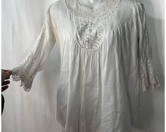 May include: White cotton blouse with a lace trim around the neckline and sleeves. The blouse has a gathered bodice and a round embroidered design in the center.