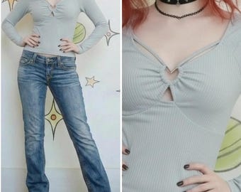 Könnte beinhalten: Hellgraues Langarm-Top mit Schlüsselloch-Detail und Ringakzent. Das Top wird mit blauen Jeans kombiniert. Das Outfit wird durch eine schwarze Choker-Halskette ergänzt.