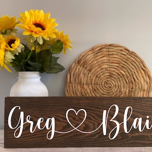 May include: A wooden sign with the names "Greg" and "Blair" written in white cursive script, connected by a heart symbol. The sign is brown and has a wood grain texture. A bouquet of yellow sunflowers is in a white vase in front of the sign.