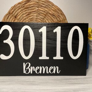 May include: Black wooden sign with white lettering. The sign reads "30110 Bremen".