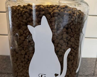 May include: A clear glass jar filled with brown pet food. A white cat silhouette is on the front, with the name "Finn" and a heart. The jar has a glass lid and is designed for pet food storage.