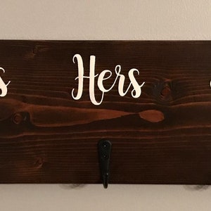 May include: A dark brown wooden sign with three hooks and the words "His" and "Hers" in white lettering. A white paw print is on the right side of the sign.