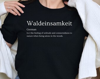 May include: A black t-shirt with white text that reads "Waldeinsamkeit" and a definition of the word in German. The definition is "(n.) the feeling of solitude and connectedness to nature when being alone in the woods."