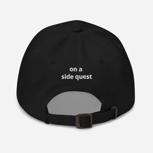 Puede incluir: Gorra de béisbol negra con el texto "on a side quest" en blanco. La gorra tiene una visera curva y una correa ajustable en la parte trasera. La gorra está hecha de un material suave.