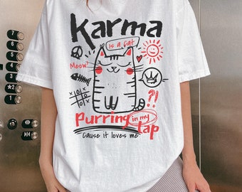 May include: White t-shirt with a graphic of a cat and the words "Karma is a cat" and "Purring in my lap". The design includes a sun, heart, crown, and other doodles in black and red.