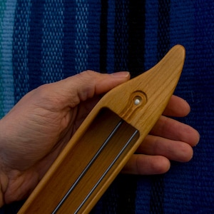 Op de afbeelding: Een houten weefspoel, vastgehouden in een hand, tegen een blauw en blauwgroene geweven achtergrond. De spoel is van lichtgekleurd hout met een afgerond uiteinde en een rechthoekige opening. Twee metalen staven zijn zichtbaar.